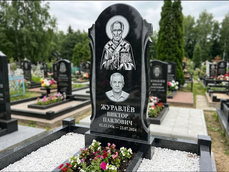 Памятник с гравировкой иконы: стела с образом в полный рост на обороте — гравировка образа Матроны Московской, низкие кусты спиреи в цвету, работа «Лебедева» в Казани
